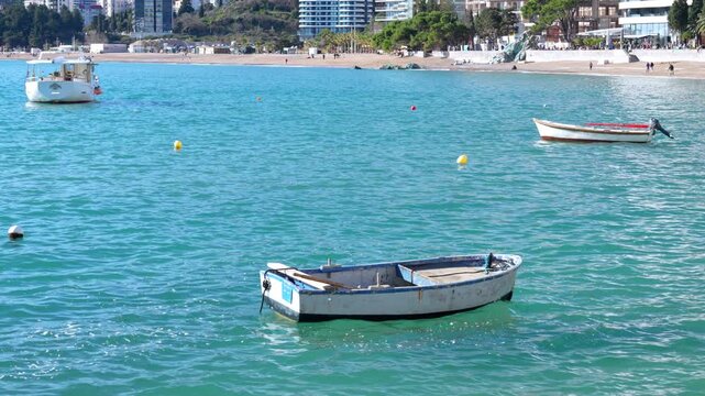 A weathered rowboat bobs gently on rippling turquoise water in a bay, with moored boats, yellow buoys, a sandy beach and seaside buildings along the distant coast.