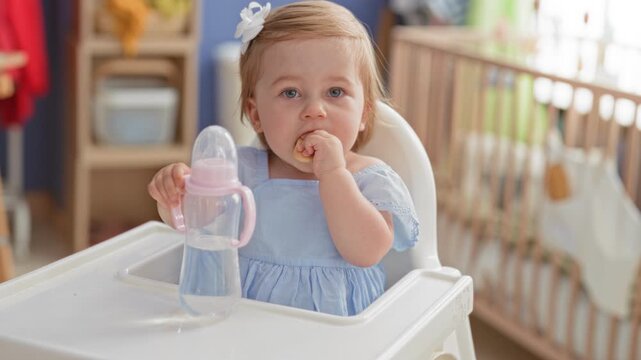 Toddler girl with blonde bow holds bottle and eats a cookie in a highchair inside a building; meal time curiosity.