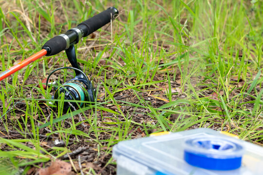 Horizontal close-up of fishing rod with spinning reel and tackle box on grass
