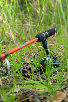 Vertical close-up of spinning reel and orange fishing rod on green grass