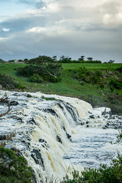 High angle view of powerful, wide waterfall cascading over dark rock formations, beneath a dramatic, cloud-filled sky, surrounded by lush green countryside, Hopkins Falls, Cudgee, Victoria, Australia
