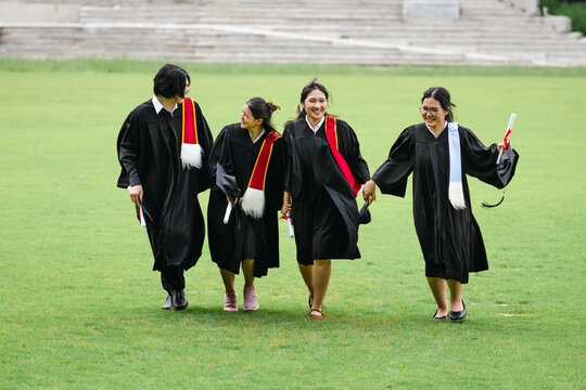 Happy group of asian university graduates running together on green lawn campus field, Cheerful graduates in academic gowns holding diplomas and hand in hand walking outdoors