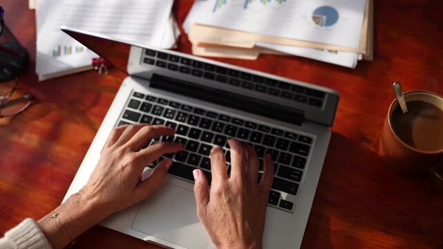 Woman typing on laptop keyboard at wooden office desk overhead view with financial documents and coffee cup for remote work concept