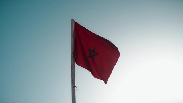 A red Moroccan flag with a green pentagram waves in the wind on a weathered pole, viewed from below against a clear sky.