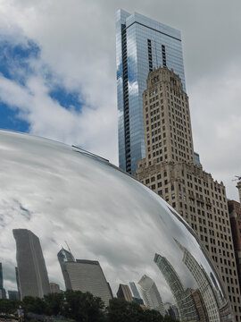 CHICAGO, IL, JUN 18, 2026: Low-angle view of Cloud Gate's mirrored surface with warped skyline reflections, beneath a blue glass skyscraper and an ornate tan limestone high-rise