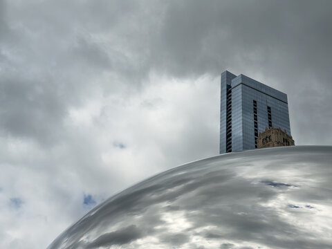 CHICAGO, IL, JUN 18, 2026: Blue glass skyscraper and an ornate historic tower peek over the top of the Bean, whose polished surface reflects gray clouds beneath an overcast sky