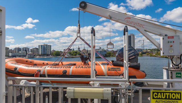 Orange safety boat aboard a ferry with davit crane, city skyline, church spires, and river visible beyond the deck railing