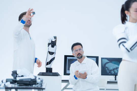 Robotics feasibility study in a busy lab, an engineer lifts the top module of the robot arm clear while a seated colleague and a bionic arm user follow the removal