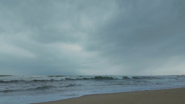 Rough turbulent sea with whitecaps rolling onto a sandy beach under heavy overcast sky, coastal city on the horizon