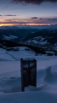Vertical video: Framing centred wooden marker post on snowy slope as sunrise lighting valley