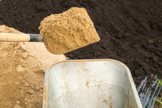A shovel full of sand is being transferred into a wheelbarrow in a construction area. Sand is scooped up with a metal shovel from the ground and placed into an empty wheelbarrow nearby.