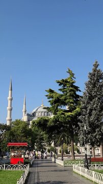 Scenic view of Sultan Ahmed Mosque in Istanbul, Turkey, framed by park trees, walking path, bright summer sky and a traditional simit stand. Travel, culture and architecture background.