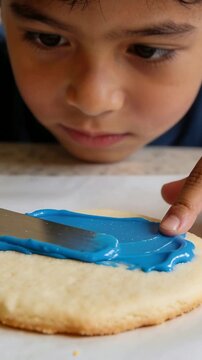 Vertical video: Placing spatula boy in blue tee laying blue icing on big cookie at home decorating