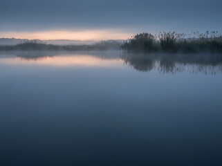 Obraz premium Distant reeds and thin fog over still blue water at pre-dawn with clean space below