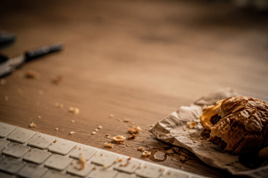 A partially eaten croissant sits on a dark brown wooden desk, scattering crumbs across the surface. Pastry crumbs on the keyboard
