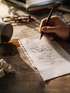 A man sketches a chaotic drawing with a black pen on a warm, honey-colored wooden table.