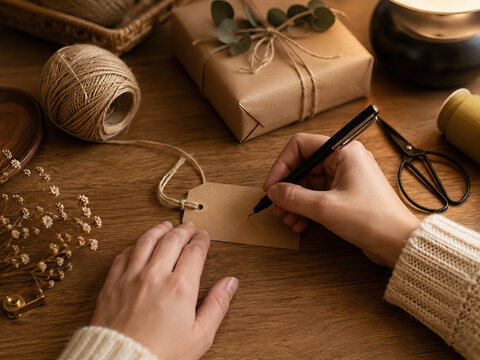 A woman meticulously writes on a brown gift tag with twine, illuminated by warm lighting on a dark wooden table.