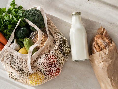 A woven mesh shopping bag overflows with fresh produce &ndash; broccoli, carrots, zucchini and parsley &ndash; on a light grey concrete surface.