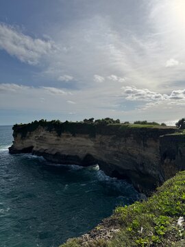 Dramatic limestone sea cliff topped with green grass and shrubs overlooks a natural rock arch where ocean waves crash below, with clear blue sea stretching to the horizon under a partly cloudy sky. Lu
