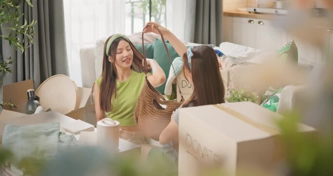 Two Asian women sit together in a cozy living room, joyfully sorting unused clothes to donate at home. Kindness, charity, promoting sustainability, generosity, eco-friendly and clutter-free lifestyle.