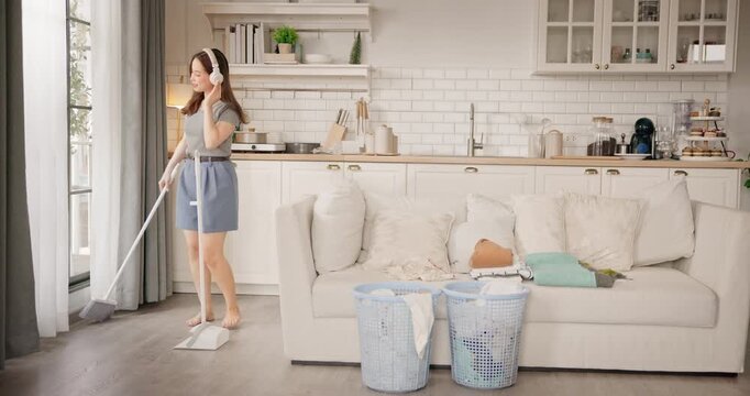 Happy young girl enjoy playful moment doing housework tidying up home on weekend. Asian women listening to music with headphones dancing and singing while cleaning floor with mop. Domestic chores.