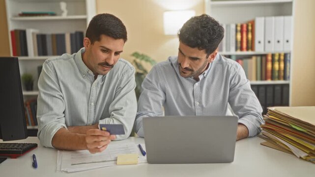 Two men holding creditcard and checking a laptop with paperwork and folders on desk in a building business setting; collaboration focus.