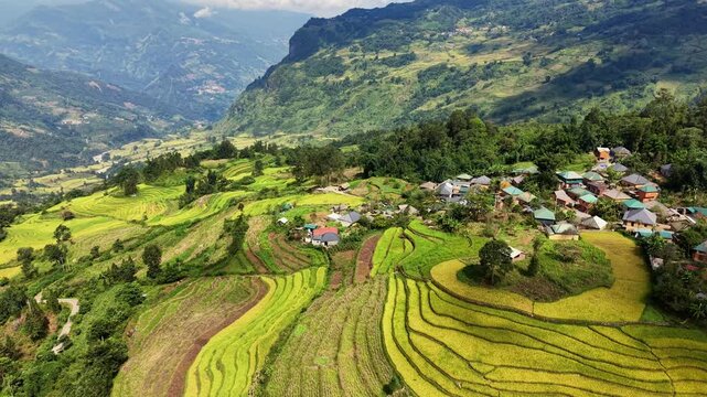 Lateral aerial view of a hilltop Ha Nhi village above green and golden rice terraces with distant mountains in Y Ty