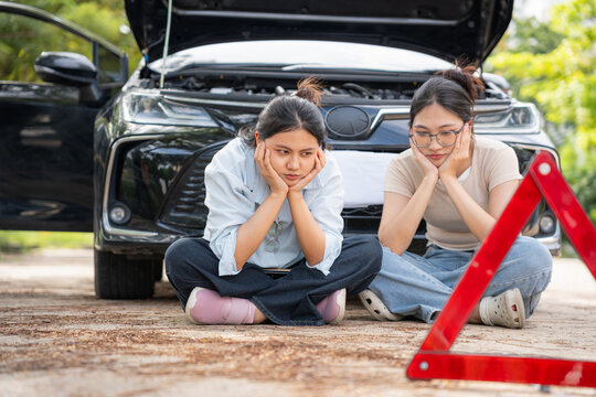 Stressed asian women sitting on ground waiting for assistance near broken down car, Worried female friends sitting in front of broken car behind emergency warning triangle