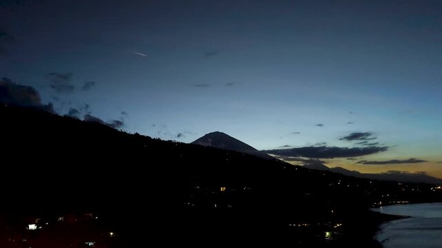 Aerial timelapse overlooking coastal and ocean landscape of Amed in Bali Indonesia with airplane flying over Mt Agung volcano at night