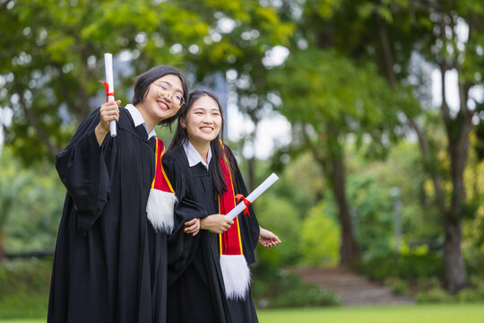 Smiling young graduates in academic gowns holding degree certificates outdoors in green park, Joyful university friends walking together holding diploma scrolls on graduation day
