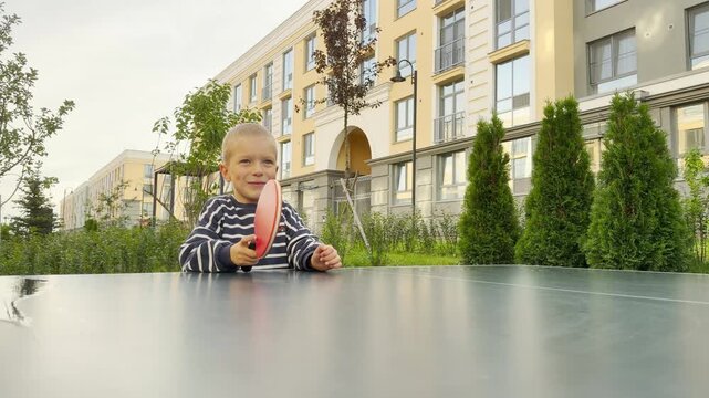 A cheerful young boy holds a red ping pong paddle with a happy smile. He pauses during a fun game at an outdoor table. Modern apartment buildings and green trees frame the pleasant courtyard.