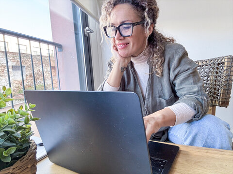 Woman smiling and focusing on a laptop, enjoying a flexible workday from a comfortable home office setup next to a window, embracing remote work and technology