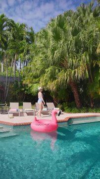 Vertical Screen: Woman relaxing by the pool walks barefoot past four empty sun loungers with rolled towels. Pink flamingo float on turquoise water, tall palm trees and blue sky on a sunny summer day.