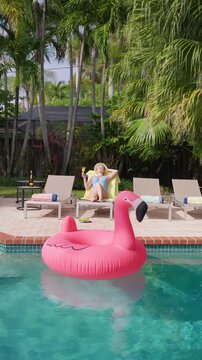 Vertical Screen: Woman relaxing by the pool in sun hat and cat eye sunglasses raises a glass of orange juice on a yellow towel. Pink flamingo float on turquoise water. Palm trees and empty loungers.