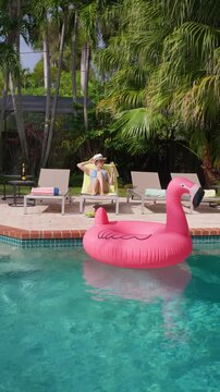 Vertical Screen: Woman relaxing by the pool holds her straw hat and a cocktail on a yellow towel. Pink flamingo float on turquoise water. Palm trees and grey sun loungers in a sunny tropical backyard.