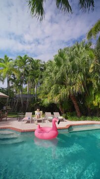 Vertical Screen: Woman relaxing by the pool on a yellow lounger with a drink. Pink flamingo float on turquoise water. Empty loungers, rolled towels, tall palm trees and white clouds on a sunny day.