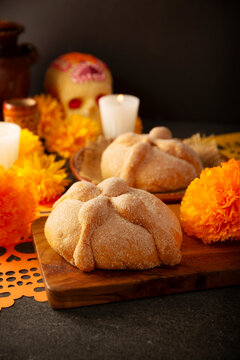 Mexican Pan de Muerto sweet bread on a wooden board, surrounded by cempasuchil marigold flowers, papel picado and candles for Day of the Dead. It is a main element in the altars and offerings.