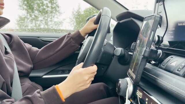 A woman holds the steering wheel while driving her car. A mounted phone displays navigation inside the dashboard. She feels focused, calm, and cautious on the quiet road.