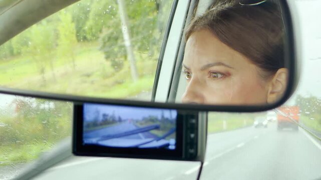 A woman's face reflects in a rearview mirror while driving near Saint Petersburg. Raindrops cover the windshield during a calm journey. The quiet drive creates a thoughtful and peaceful atmosphere.