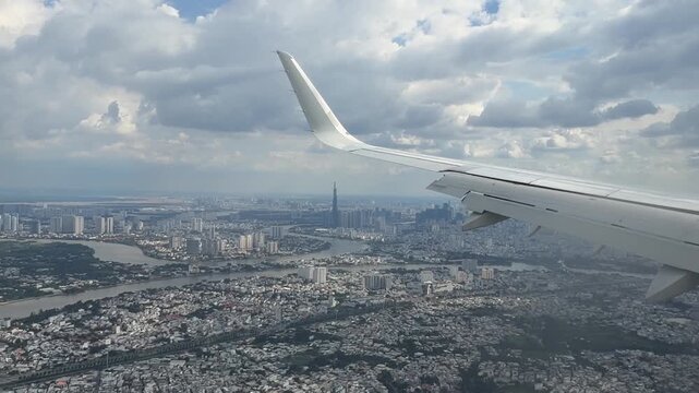 an airplane passenger window during the landing approach to Tan Son Nhat International Airport in Ho Chi Minh City, Vietnam. The sprawling urban cityscape, winding rivers, and skyscraper in cloudy day