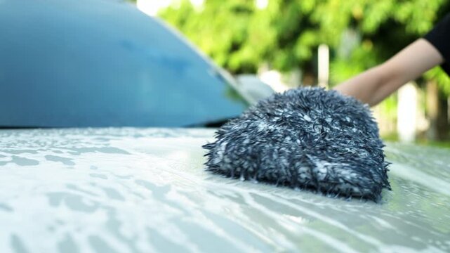 Close-up of person using microfiber wash mitt to clean soapy car hood outdoors. DIY car wash, auto detailing and vehicle maintenance lifestyle concept.