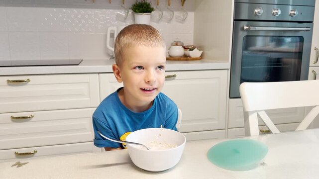 A happy young boy laughs cheerfully while enjoying his breakfast. He sits at a white table in a bright modern kitchen. His joyful smile shows pure morning delight.