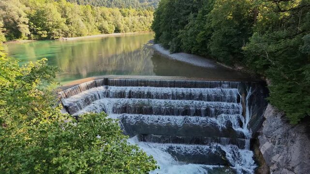 Wide view of the Lechfall, a tiered weir where the turquoise Lech River tumbles over five steps between green forest in Fussen, Bavaria, Germany, on a summer afternoon.