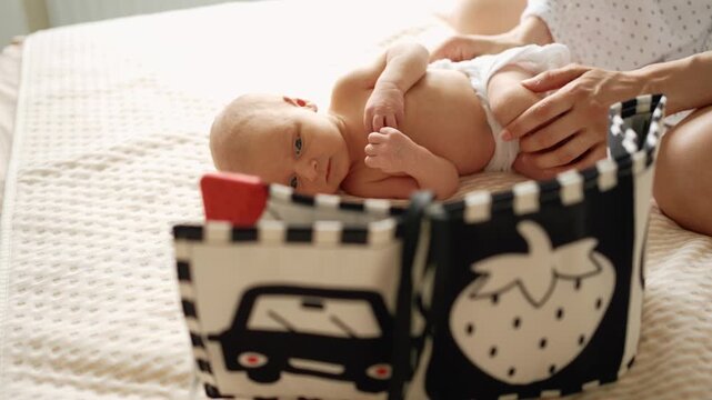 Newborn infant with blue eyes wearing a diaper lies on a soft bedspread, focusing on a black and white high contrast soft book, a vital tool for early visual development and stimulation
