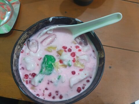 Indonesian traditional dessert Es Pisang Ijo served in a bowl with rice flour porridge, tapioca pearls,,green banana rolls sweet pink syrup, and coconut milk.