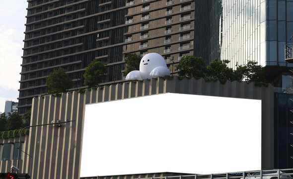 5 OCT 2026, Bankkok, Thailand. Giant white inflatable cartoon character, playful and cute, sits on rooftop garden of modern city building, above large blank billboard, urban outdoor scene, daylight