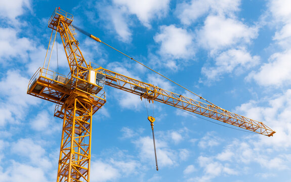 Large yellow construction crane jib extends toward blue sky with white clouds. Heavy industrial machinery uses steel lattice boom, lifting hook for building projects