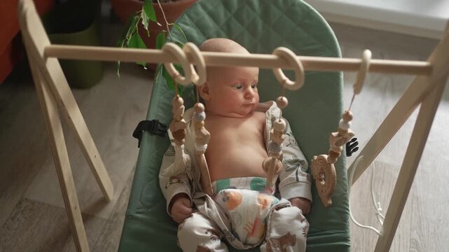Curious newborn baby boy looking at wooden sensory toys while lying in a comfortable bouncer. Infant cognitive development through play, exploring shapes and textures in a sunlit nursery room