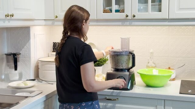 A focused woman operates a food processor in a bright kitchen. She shreds fresh vegetables with calm concentration. Her quiet work creates a peaceful and cozy mood.