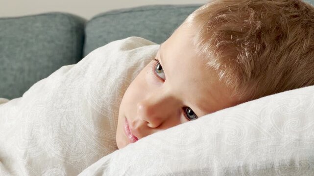 A young boy rests under a cozy blanket and watches tv. He lies peacefully on a soft pillow on the sofa. His quiet gaze shows calm, gentle comfort.